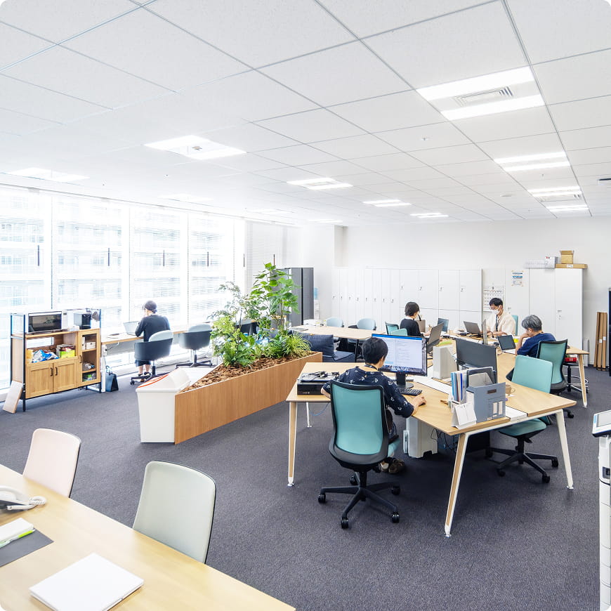 明るい自然光が差し込むオープンなオフィスで、複数の社員がデスクワークをしている