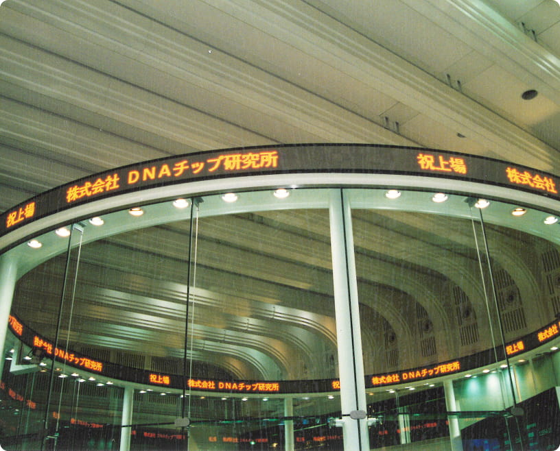 東京証券取引所内と思われる場所で、「株式会社DNAチップ研究所」の文字が電光掲示板に表示されている