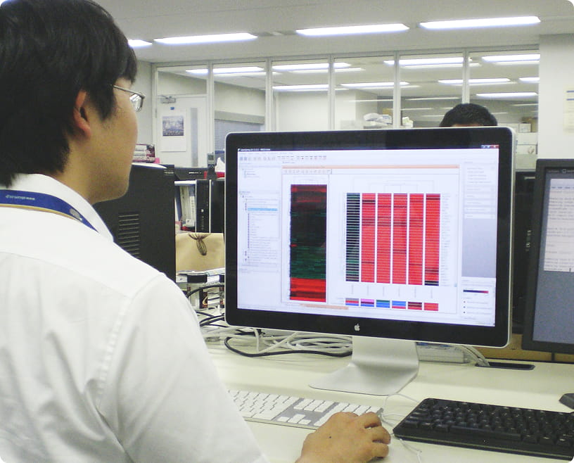 白衣を着た男性が、デスクでモニターに表示された遺伝子解析データのようなカラーマップを見ている